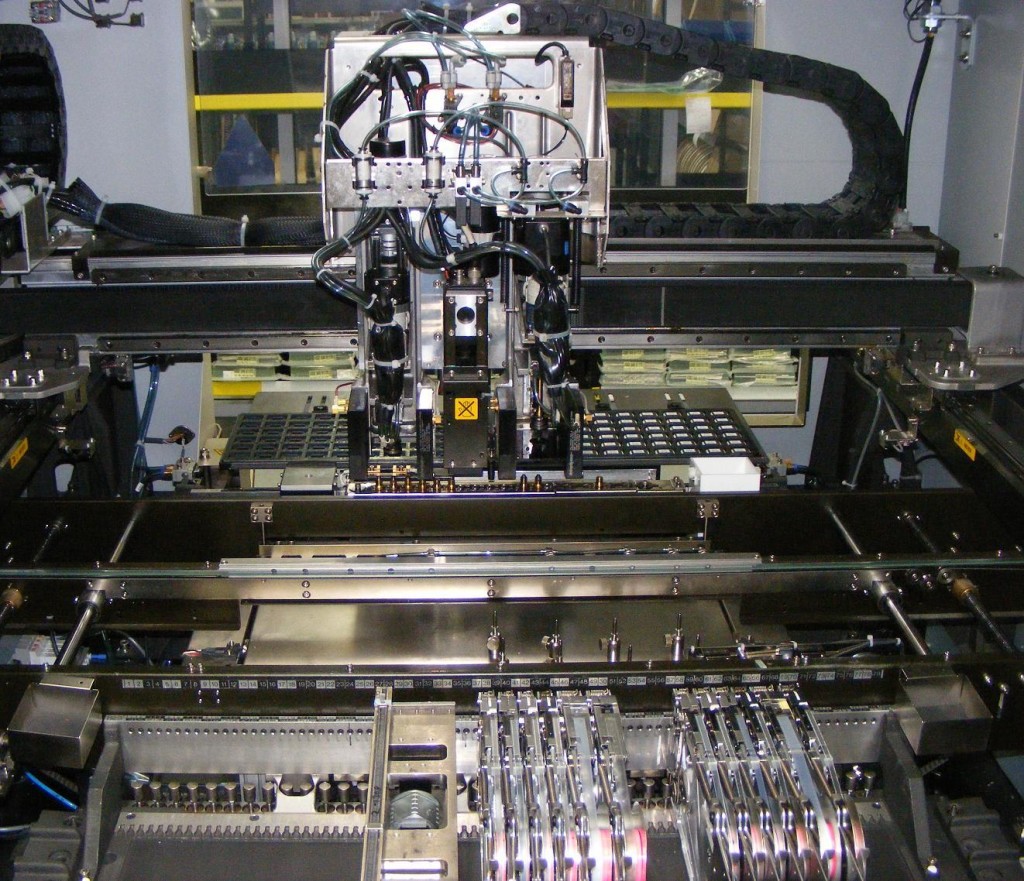 Internals of a Juki KE2010L 4 head pick and place surface mount machine. the machine is used to place electronic components that are soldered to the face of a printed circuit board rather than having leads go through holes in the board.   The machine is loaded with standard 8" reels on mechanical feeders (front right) and a tray of QFP microprocessors (rear center)  Picture by Peripitus